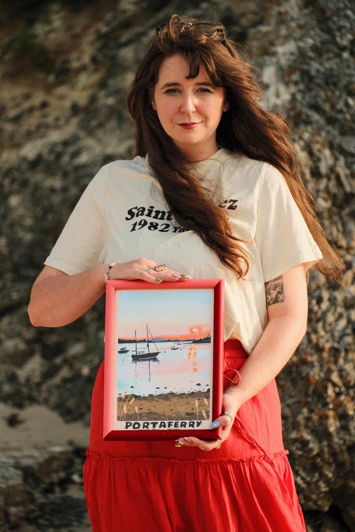 A woman with long brown hair stands outdoors against a rocky background, holding a red-framed picture that reads 'PORTAFERRY'. She is wearing a cream graphic t-shirt and a red skirt.